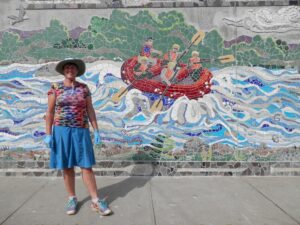 Kate Hartley, a white woman in a colorful tank top and bright blue skirt stands in front of the rafting scene in the mosaic. The sun is shining and she is smiling.