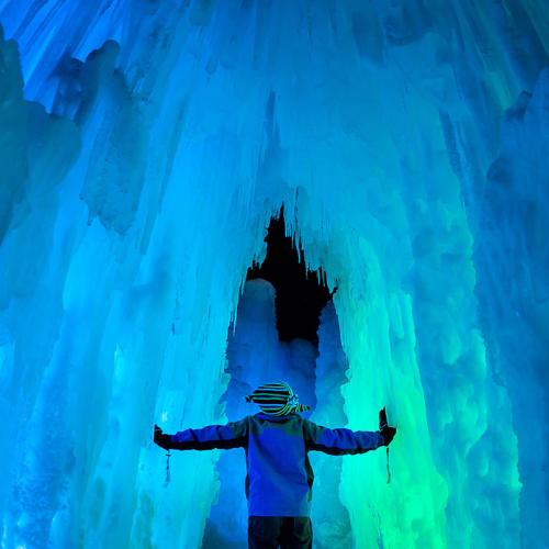 A person stands surrounded by blue and green glowing ice cavern. They stand with their back to us and their arms outstretched on either side of them, touching the walls.
