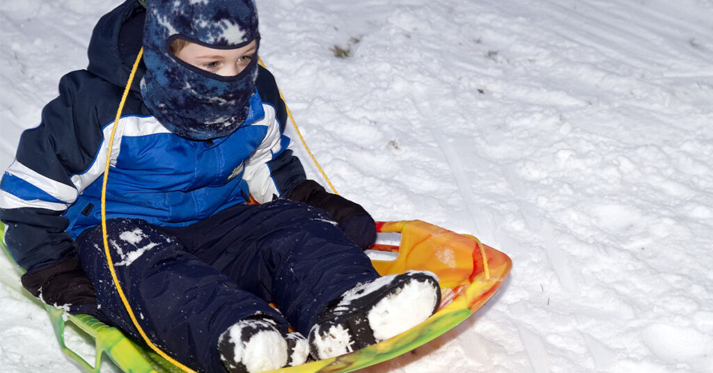 A young boy bundled up in a blue jacket and black snowpants flies down a snowy hill on a yellow sled.