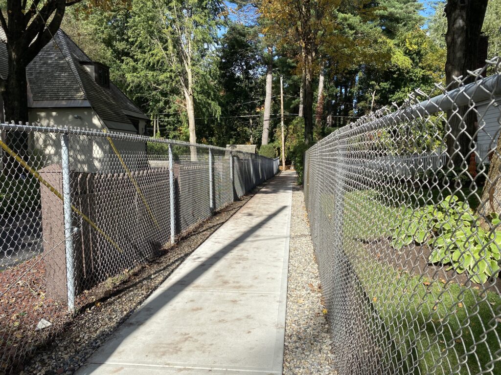 The concrete path is empty and has chainlink fence on both sides, leading to the park.
