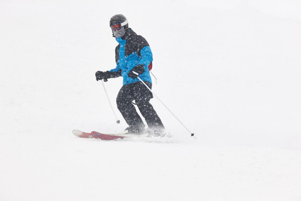 A man skis down a snowy white slope. He is wearing a black and blue ski jacket, black snow pants, and a black helmet.