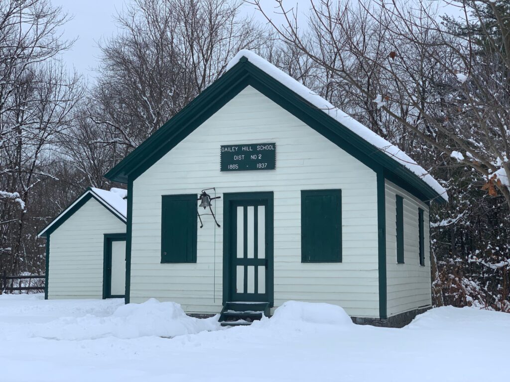 Snow covers the roof and grounds of a quaint wihte schoolhouse with green trim.